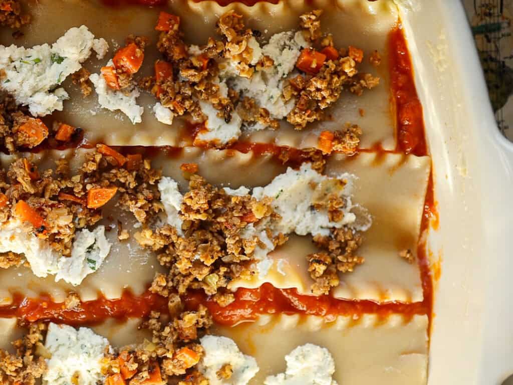 Close-up of unbaked lasagna in a white dish. Visible layers of pasta sheets, red tomato sauce, ricotta cheese, and a mix of sautéed ground vegan meat with chopped carrots.